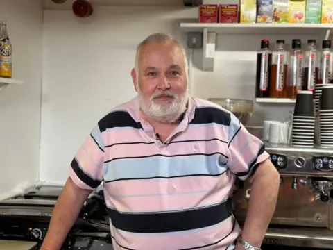 Grey-bearded Mark Breen wearing a pink striped polo shirt standing in his cafe kitchen with disposable mugs and glasses seen behind him