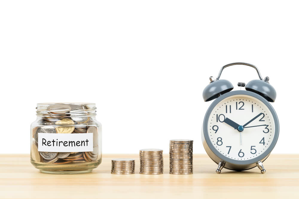 Glass jar filled with coins labeled 'retirement'; pictured next to it are 3 vertical stacks of coins growing in size from left to right and an old style alarm clock